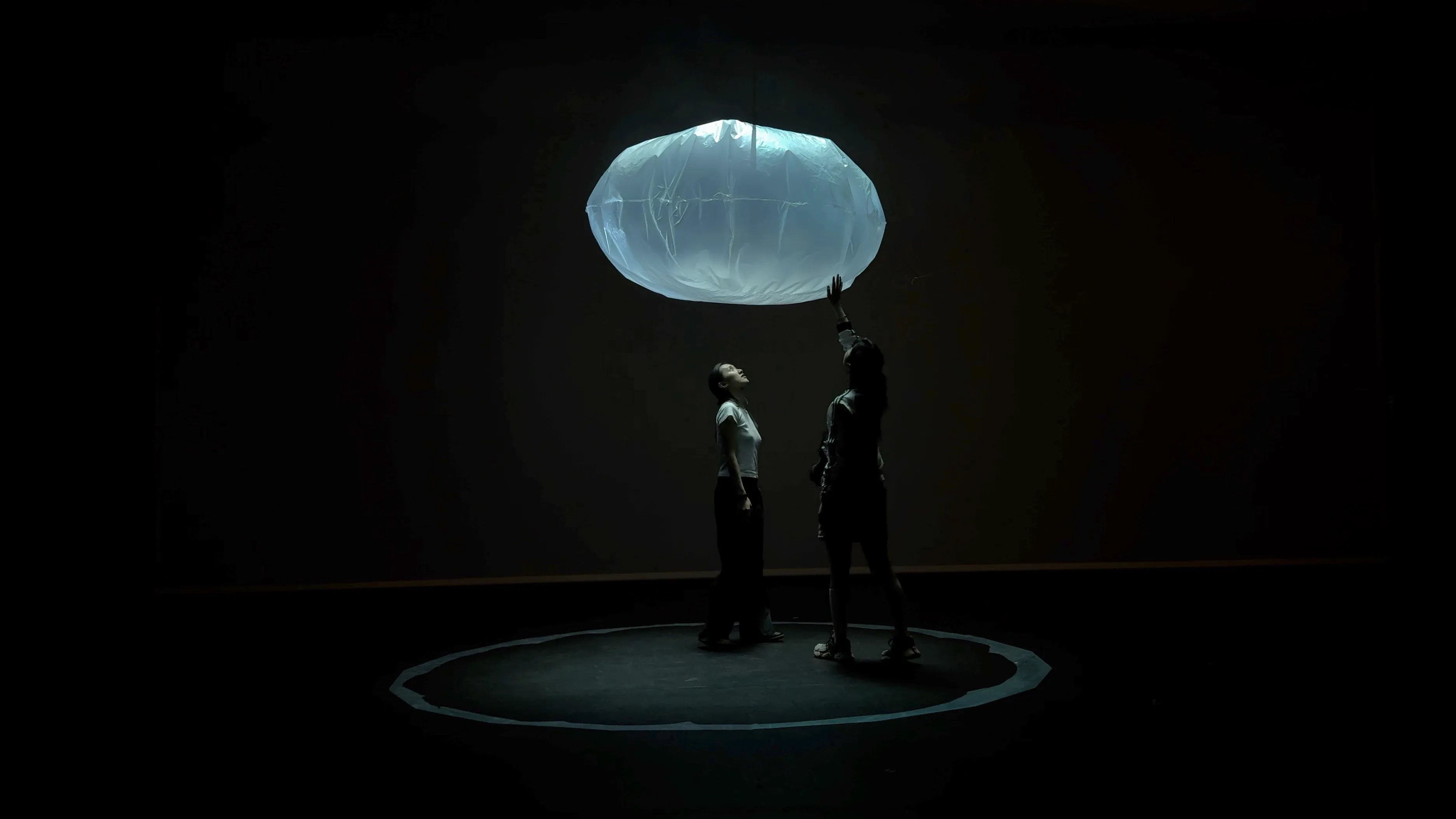 Two visitors standing beneath the floating bubble in a dark room, one reaching up to touch it